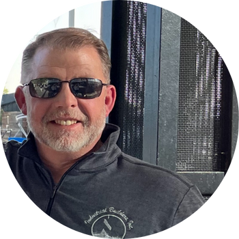 Man wearing sunglasses smiles, with a gray beard, and a black jacket that says