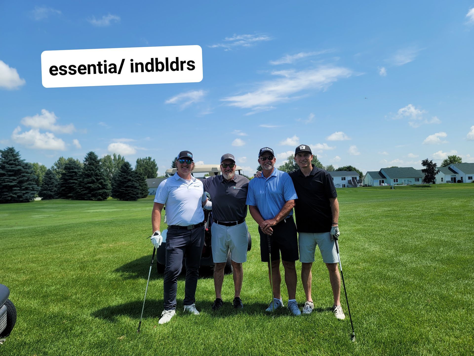 Four men in golf attire on a green field under a blue sky, posing with clubs.