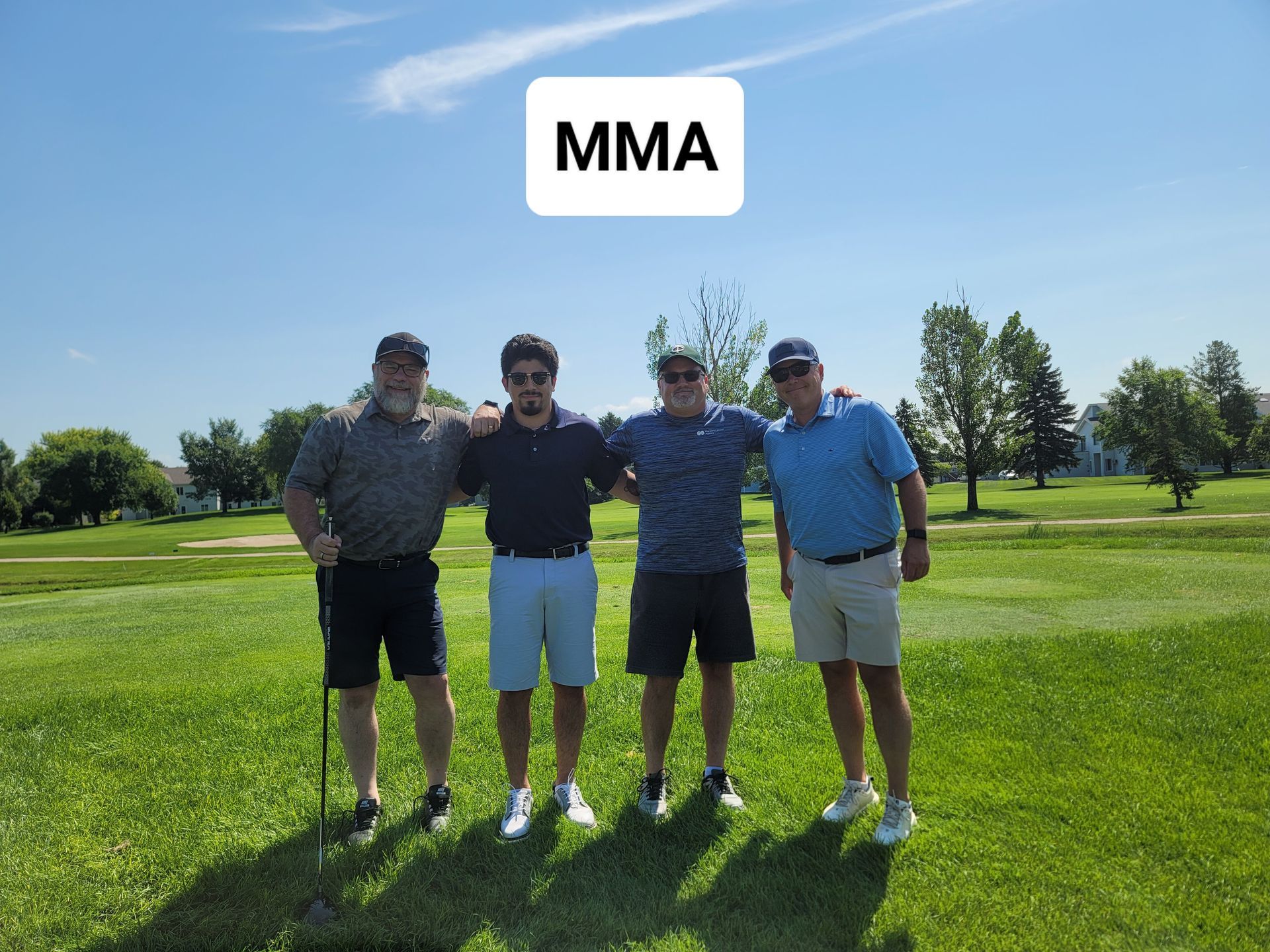 Four men standing together on a golf course under a blue sky, arms around each other.