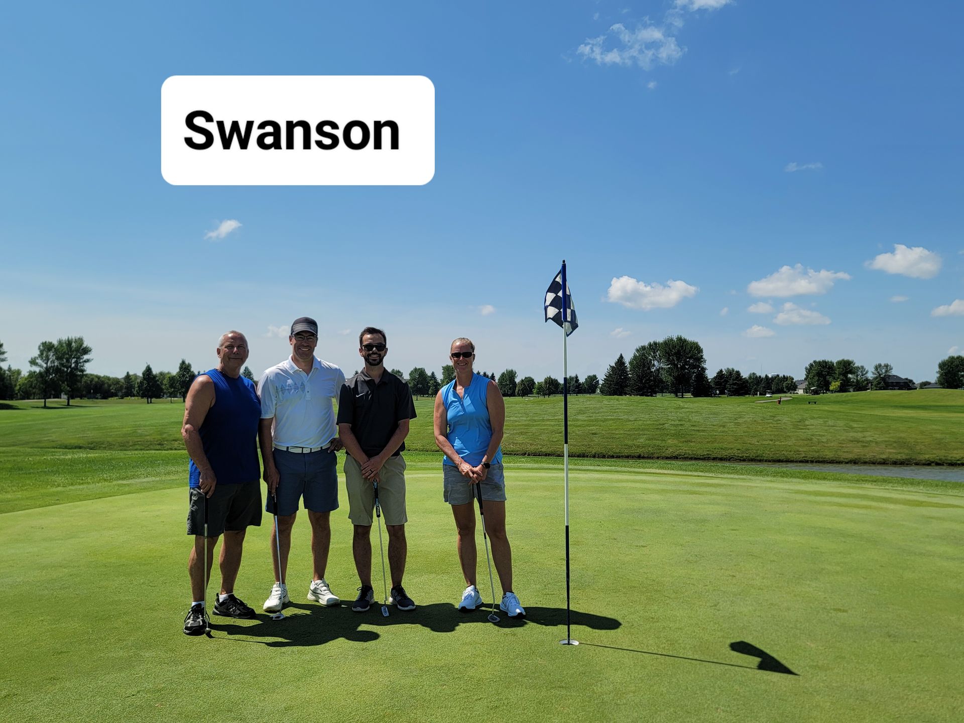 Four people standing on a golf course green under a sunny sky near the flag.