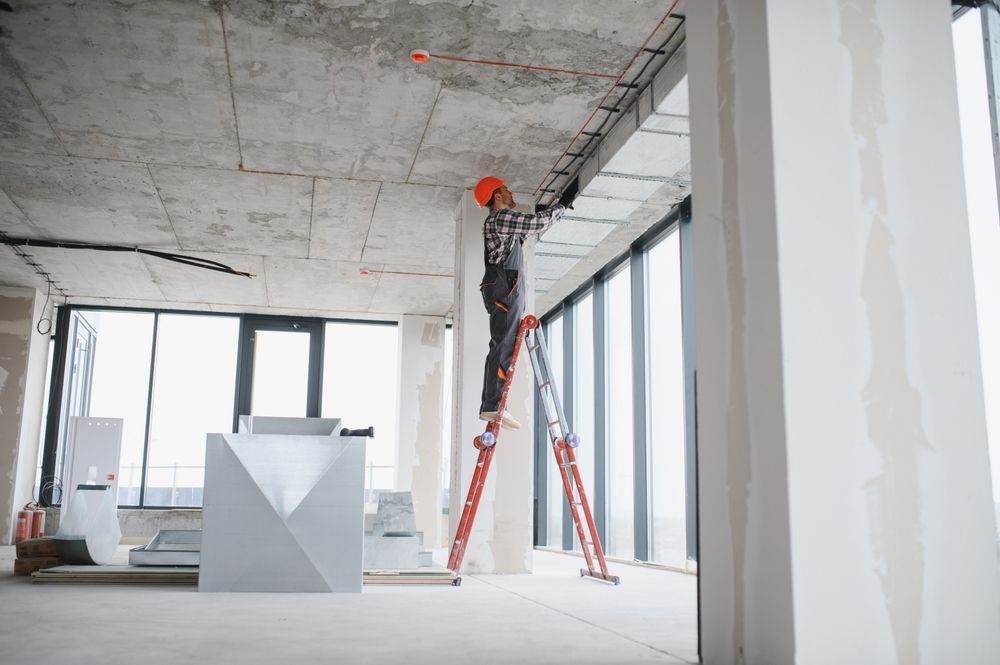 Construction worker on a ladder in an unfinished building, installing wiring on the ceiling.