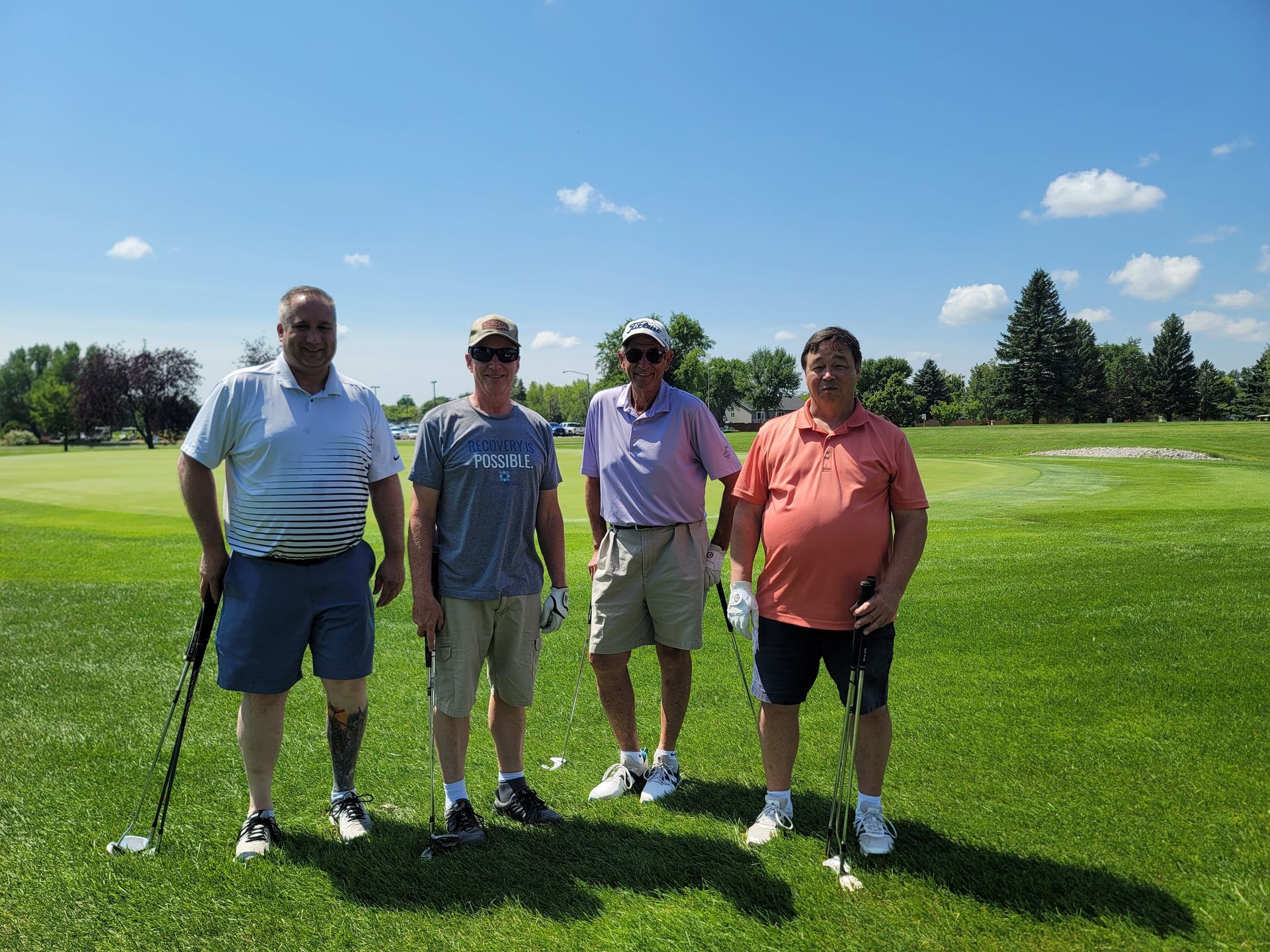 Four men on a sunny golf course, each holding a club, posing for a photo on the green.