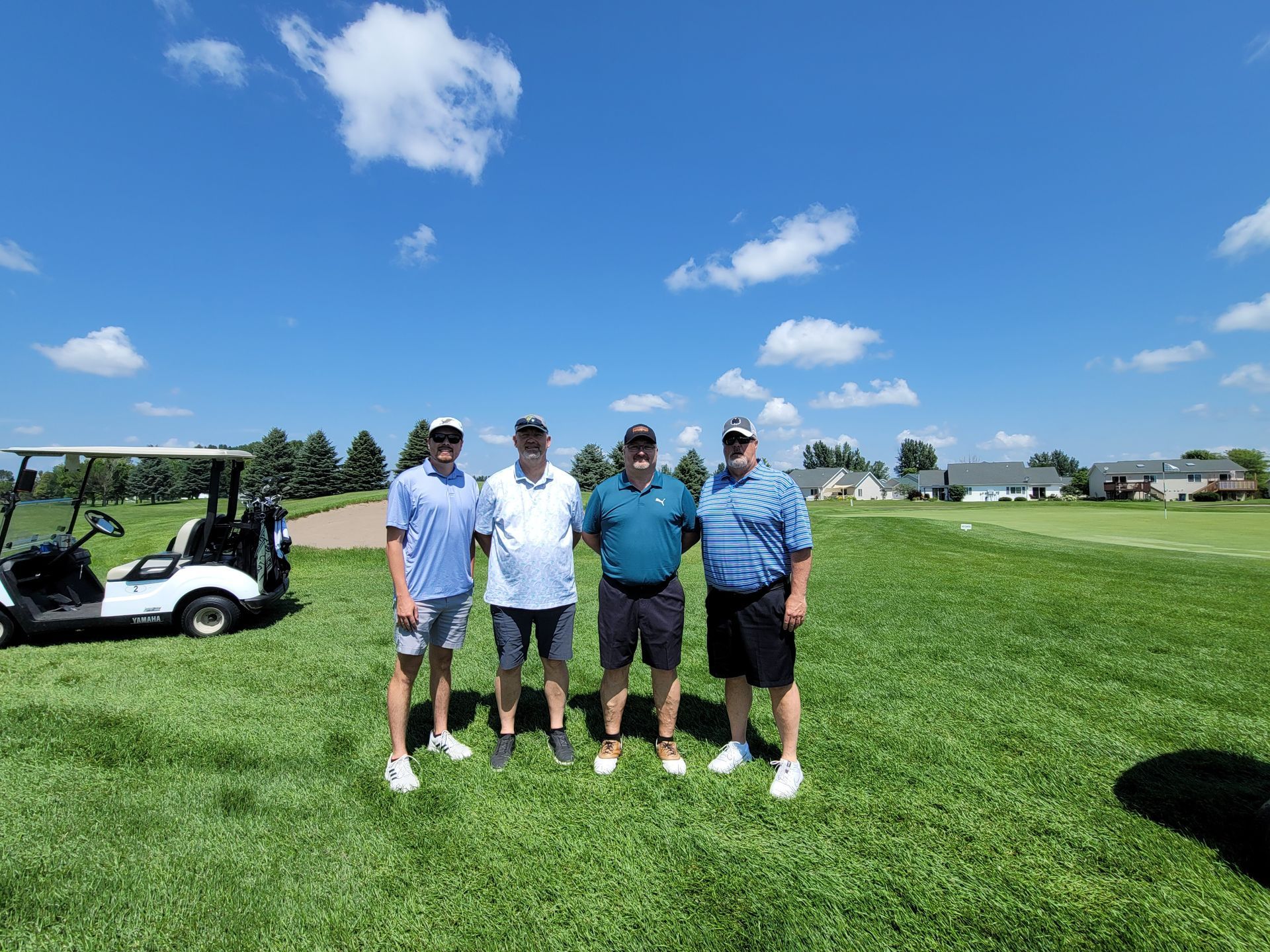Four men on a golf course pose for photo. Sunny day with golf cart nearby, green grass, and blue sky.