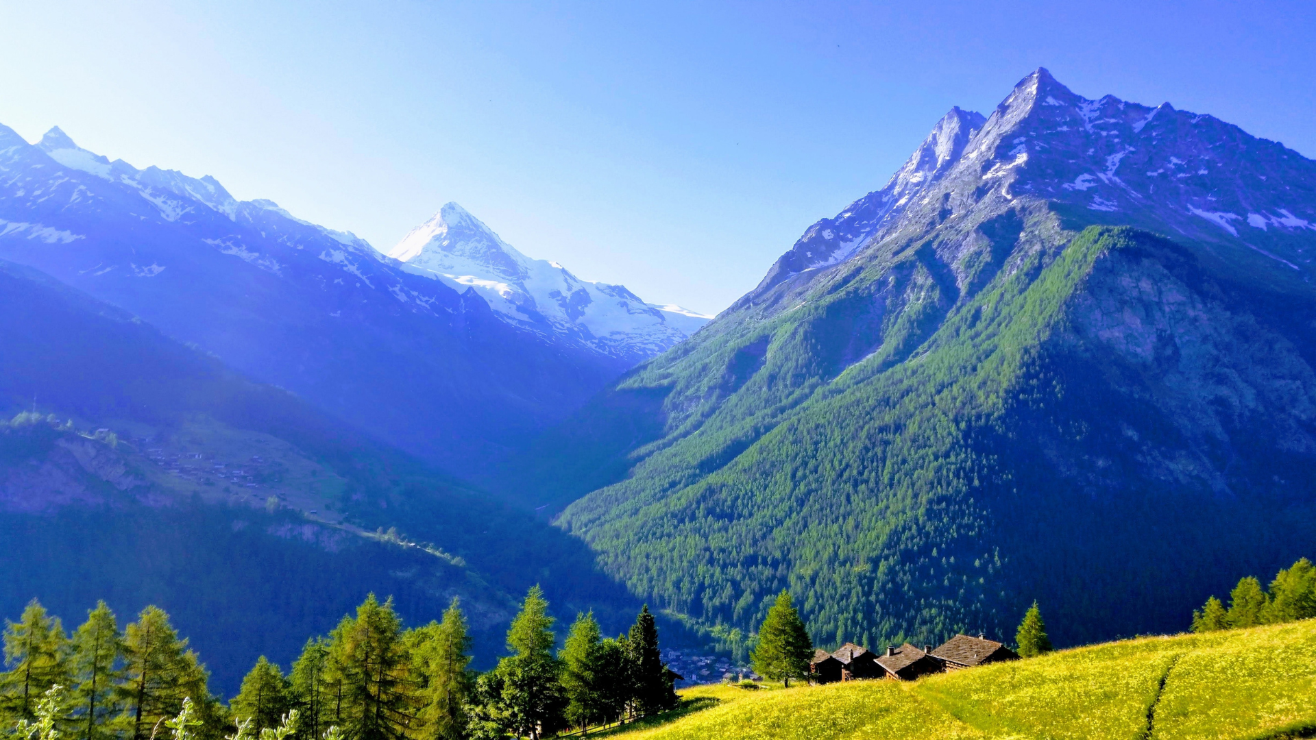 Mountains with snow-capped peaks rise above a green valley and meadow dotted with small cabins.