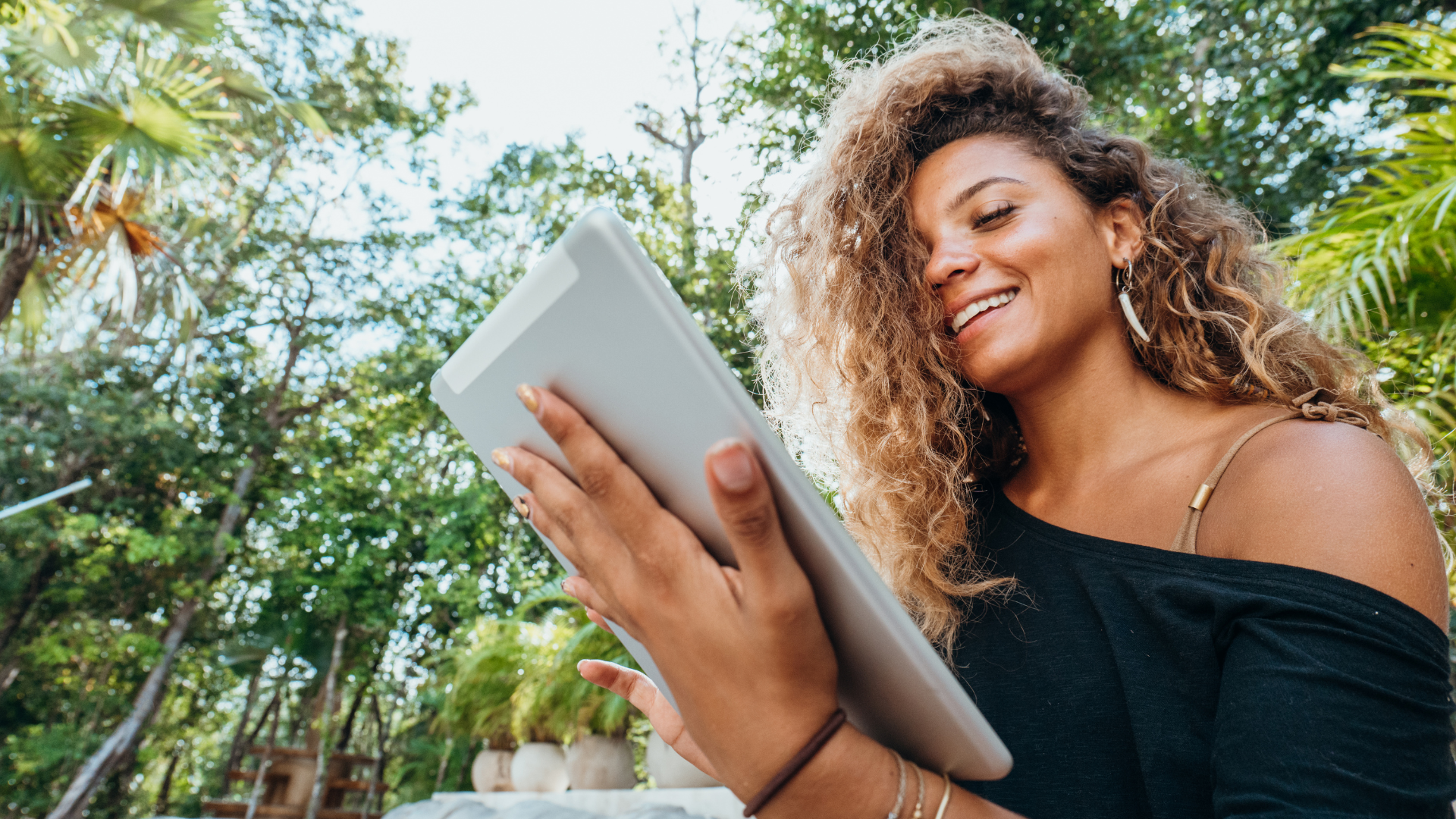 Woman with curly hair smiles while looking at a tablet outdoors, surrounded by green foliage.
