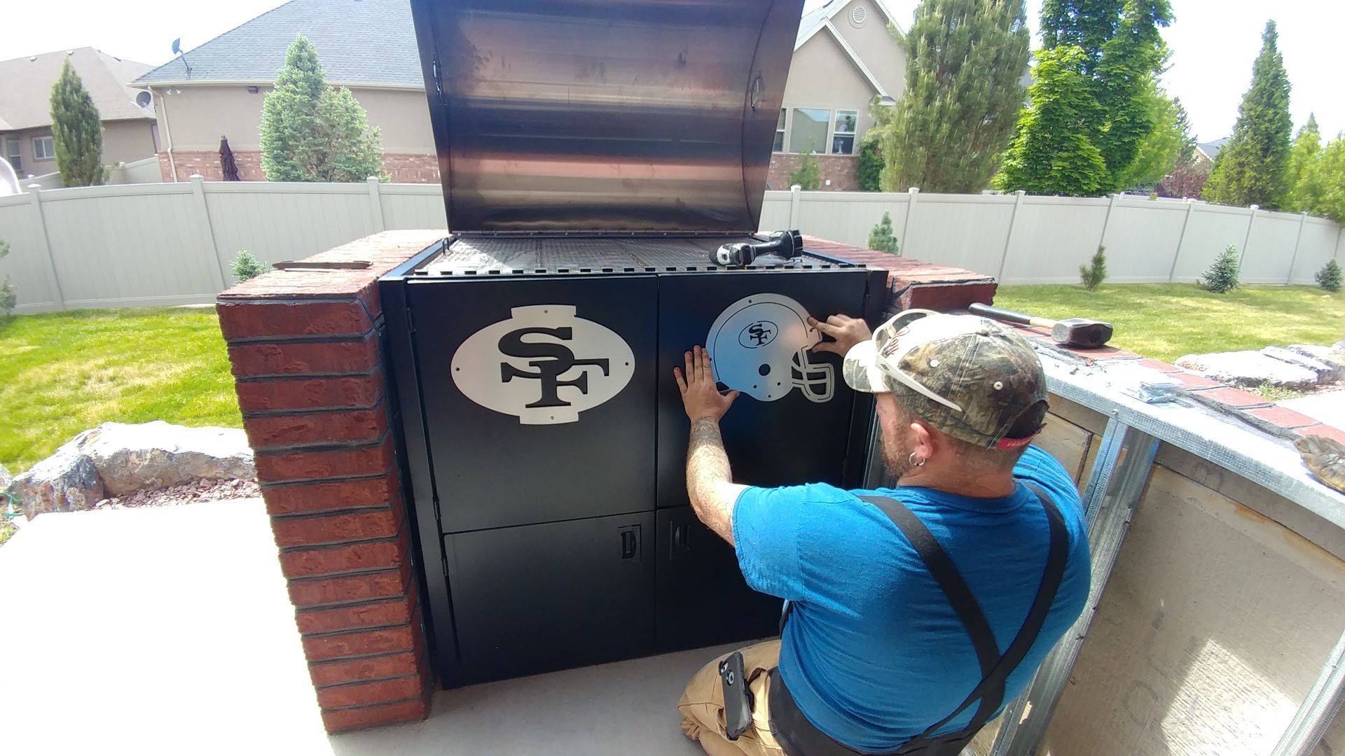 A man installing a circular gauge on a black barbecue, framed by red brick, outdoors on a sunny day.