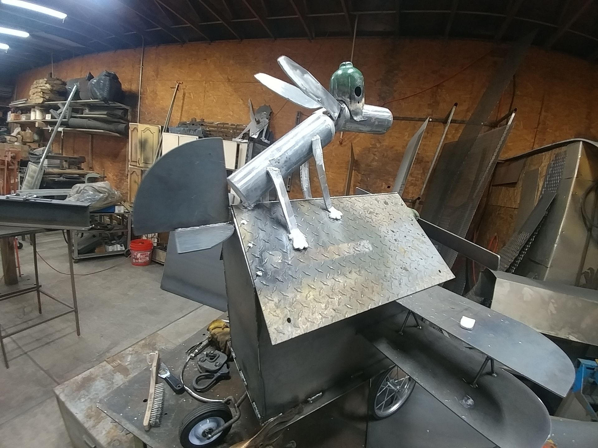 Metal sculpture of a donkey with wings atop a wheeled cart, inside a workshop.