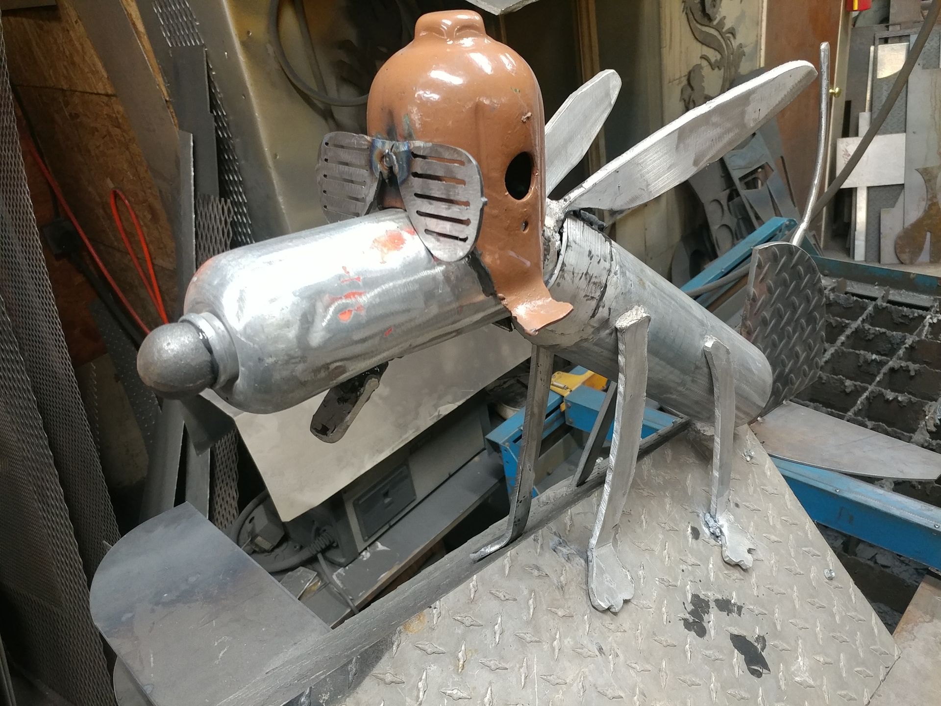 Metal sculpture of a dog-like creature with sunglasses, made from repurposed materials in a workshop setting.