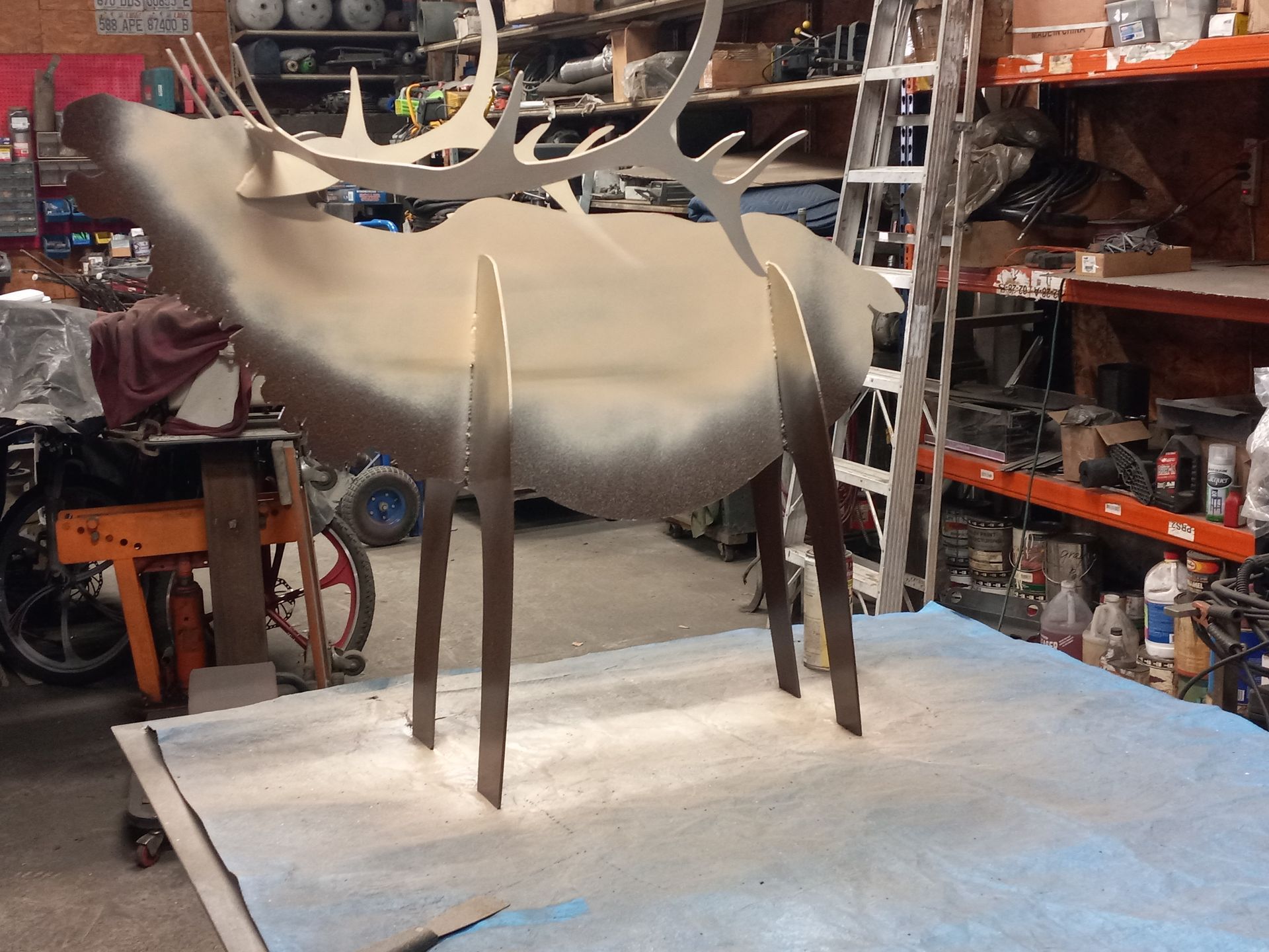 Metal elk sculpture in a workshop, painted in shades of brown and beige.