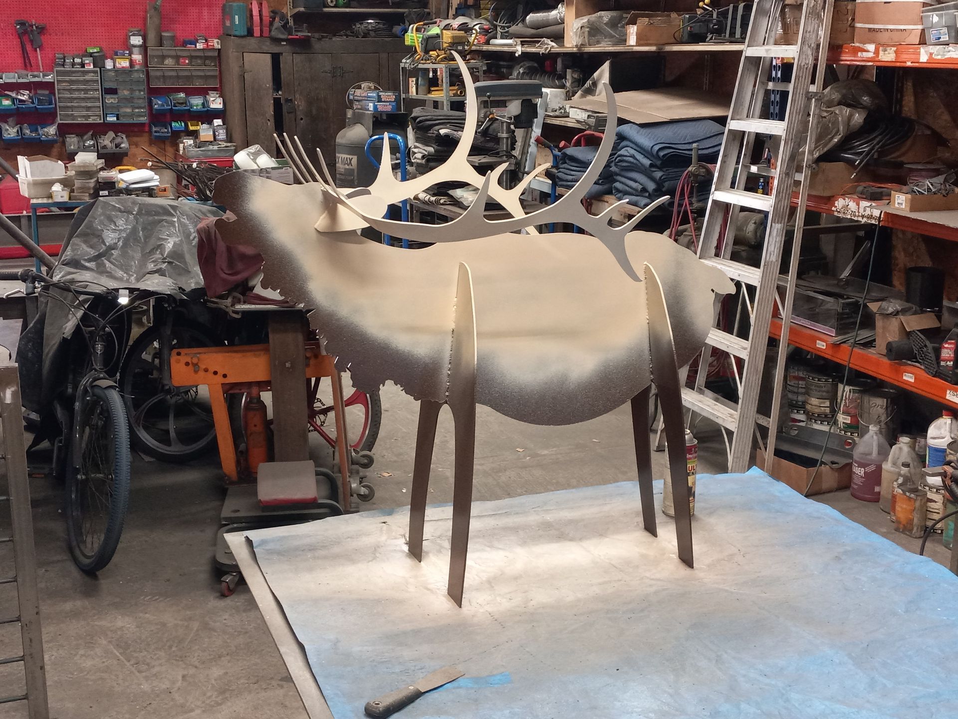 Metal sculpture of an elk in a workshop setting. The elk is beige, with long legs and large antlers.