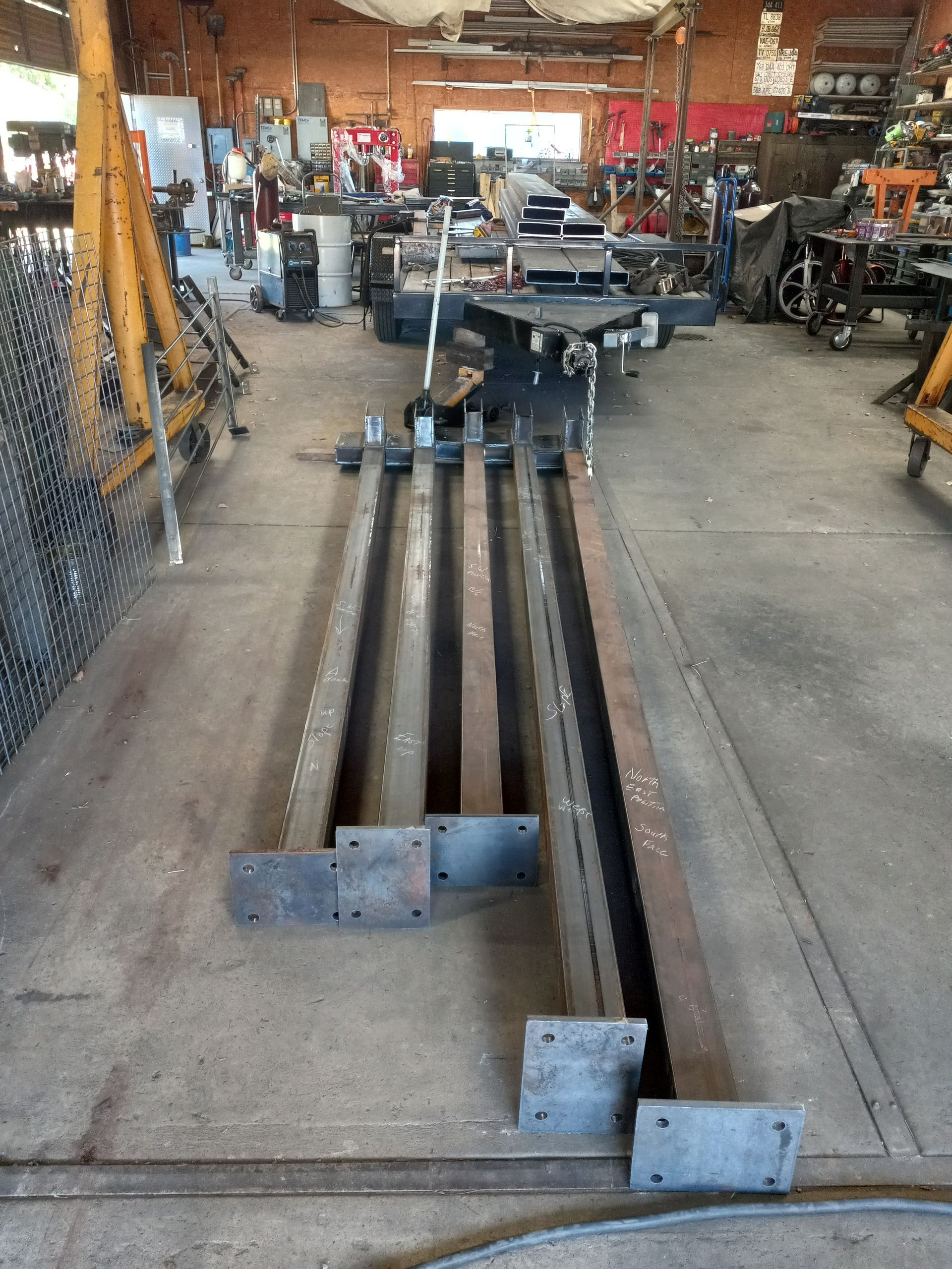 Metal fabrication shop with long steel beams, work in progress. Gray floor, various equipment visible.