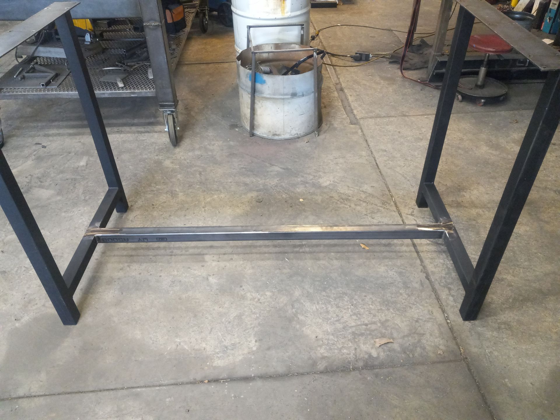 Black metal table base on concrete floor, with a crossbar. Workshop setting.