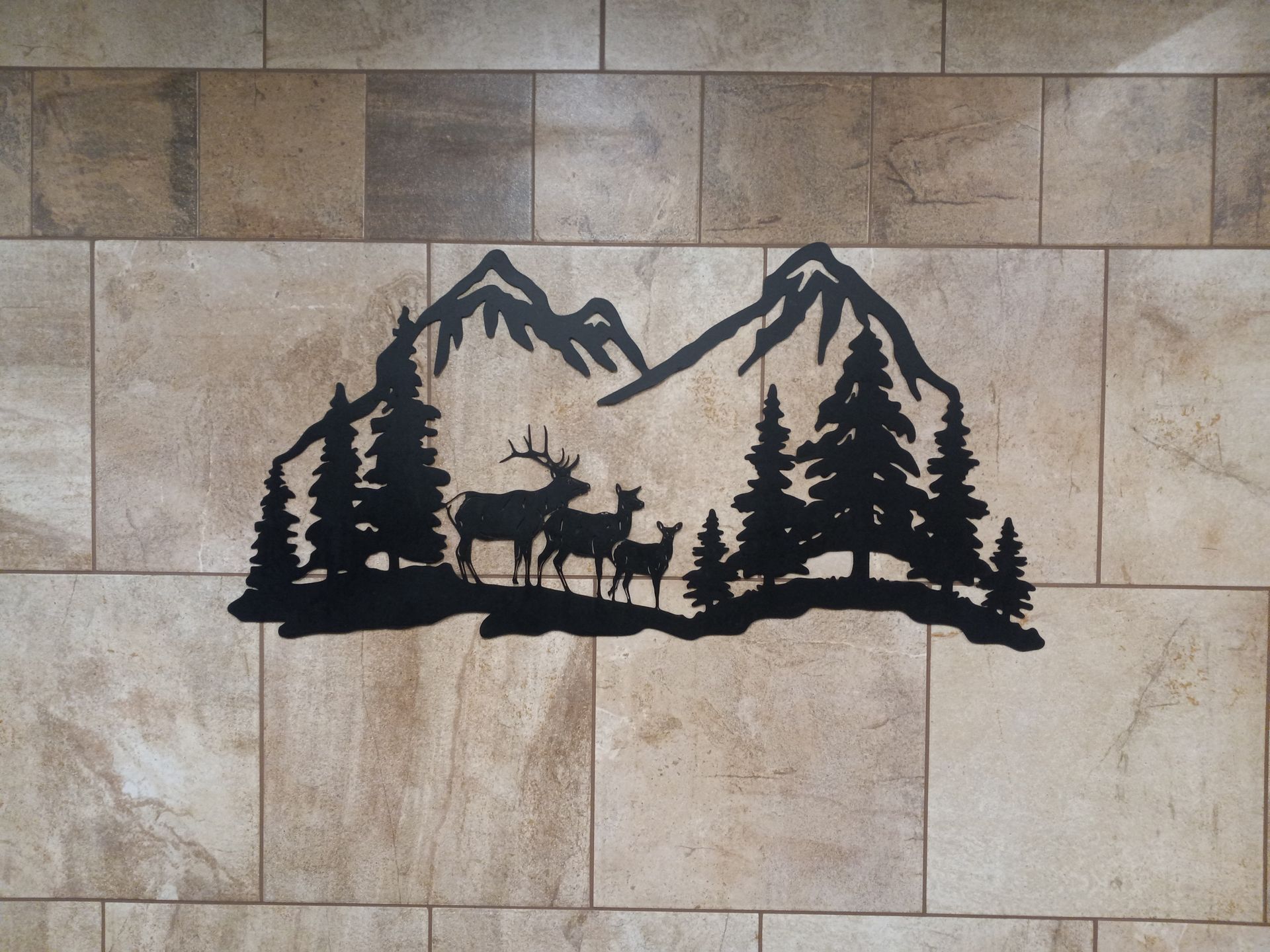 Black metal wall art of a deer family in front of trees and mountains against a tiled wall.