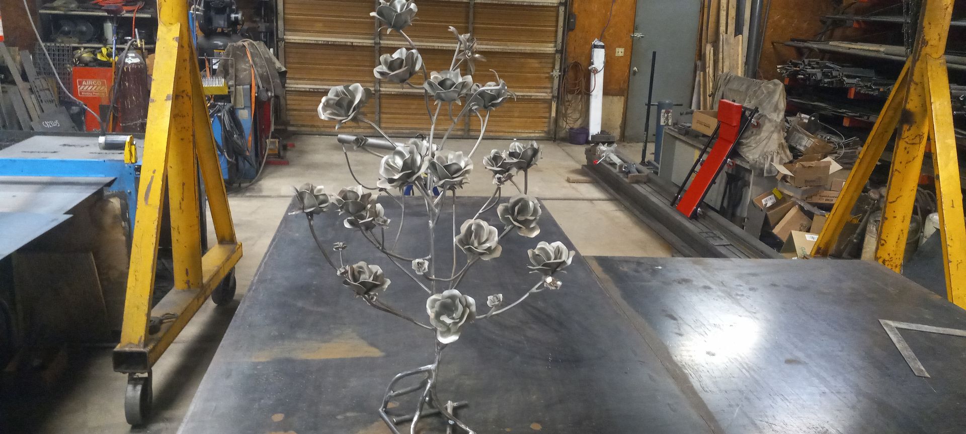 Metal sculpture of a flowering tree on a metal table, in a workshop. Yellow supports frame the table.