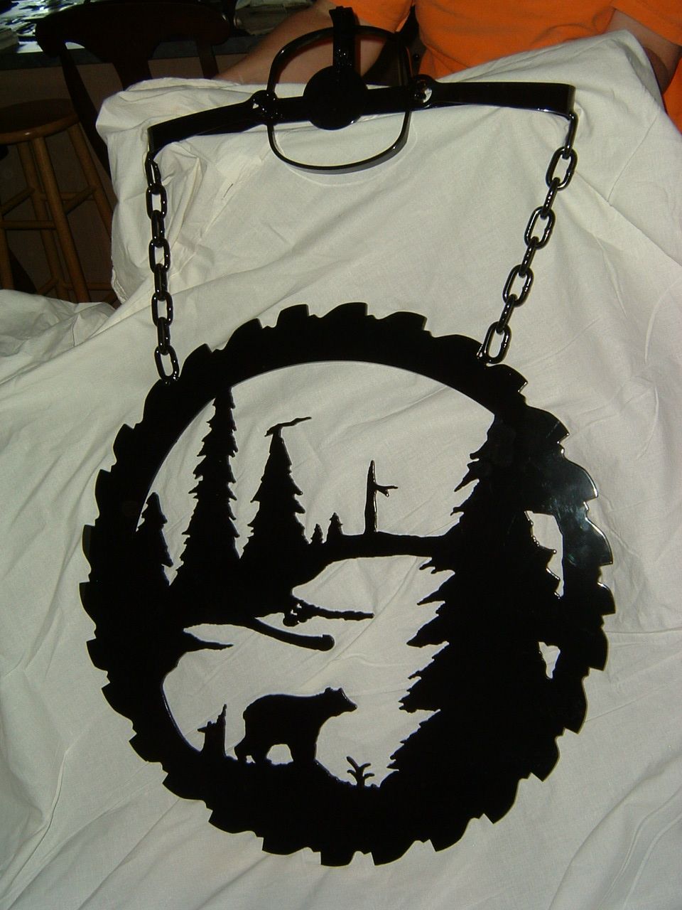 Black metal wall hanging of a forest scene cut into a circular saw blade. Bear silhouette near a lake.