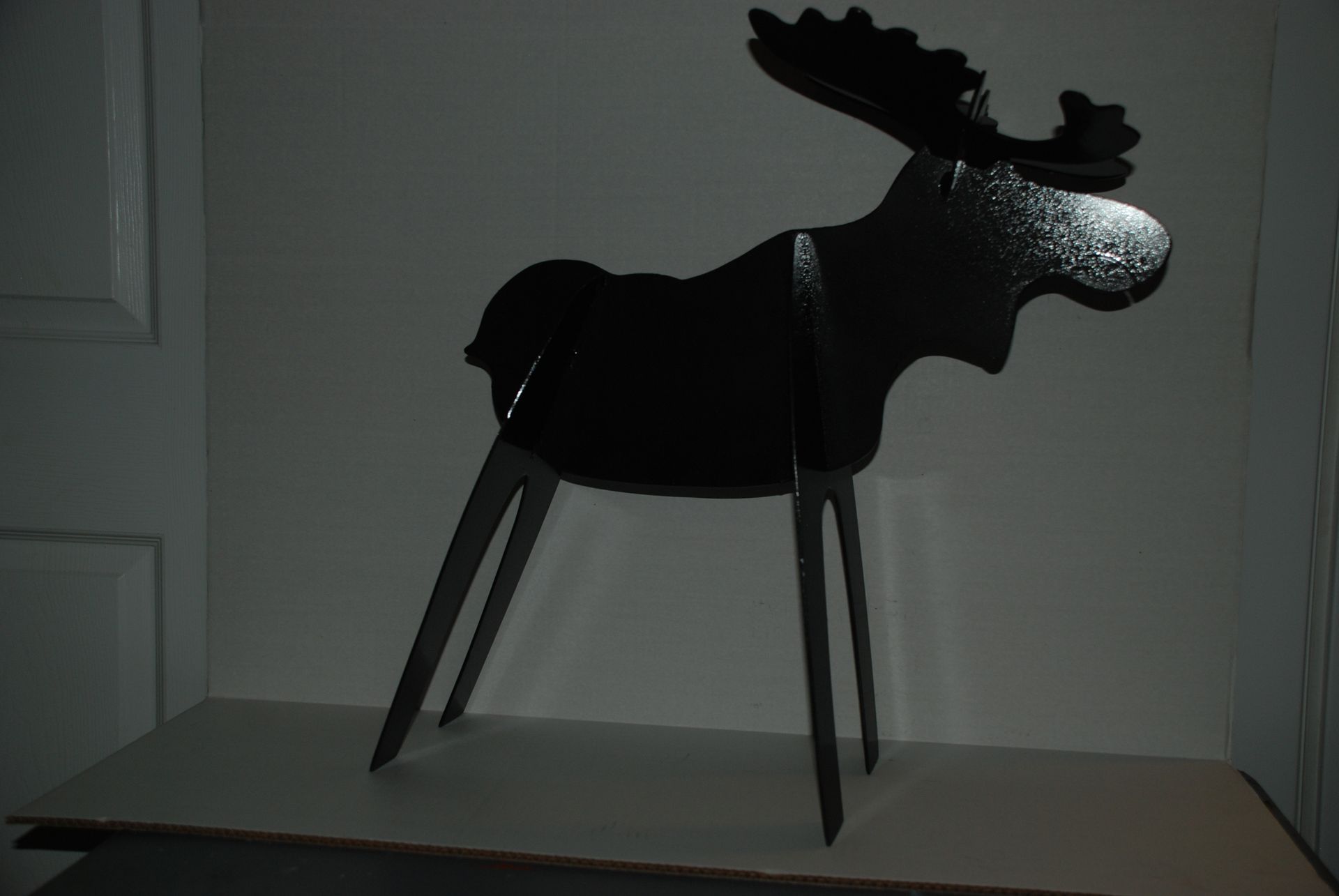 Black silhouette of a moose statue with large antlers and thin legs, standing on a light-colored surface.