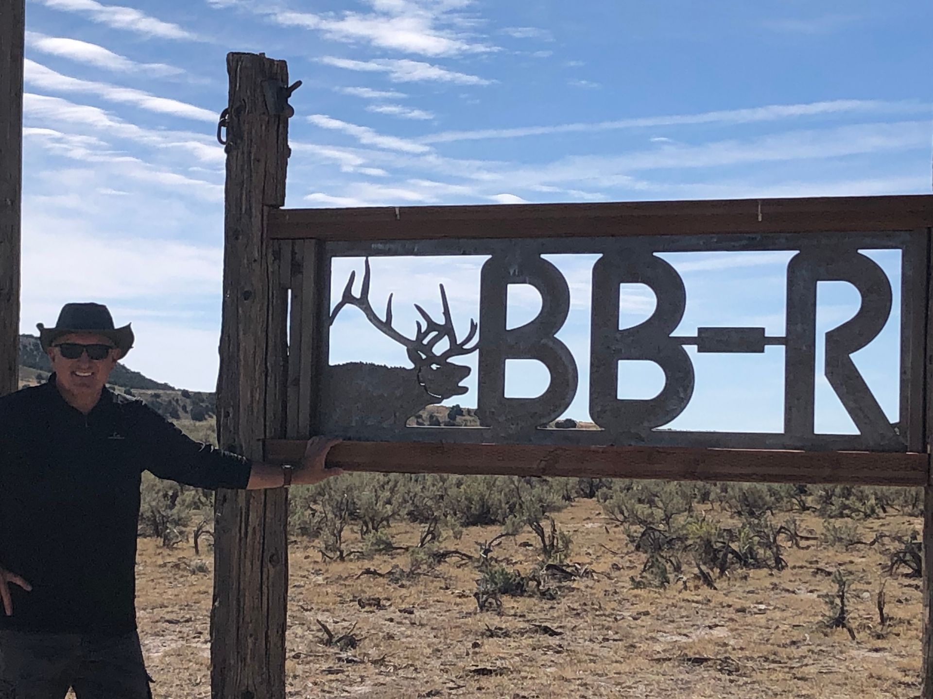 Man in a hat poses by a weathered wooden sign: BB-R with an elk silhouette. Arid landscape, blue sky.