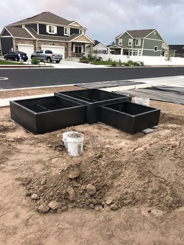 Three dark gray raised garden beds on dirt in front of new houses. White bucket in the middle.