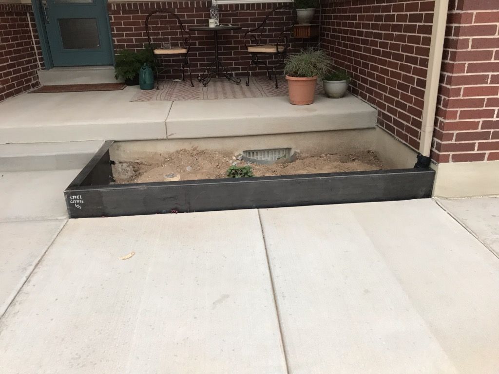 Concrete steps leading to a building's entrance. A rectangular sunken planter is in front.