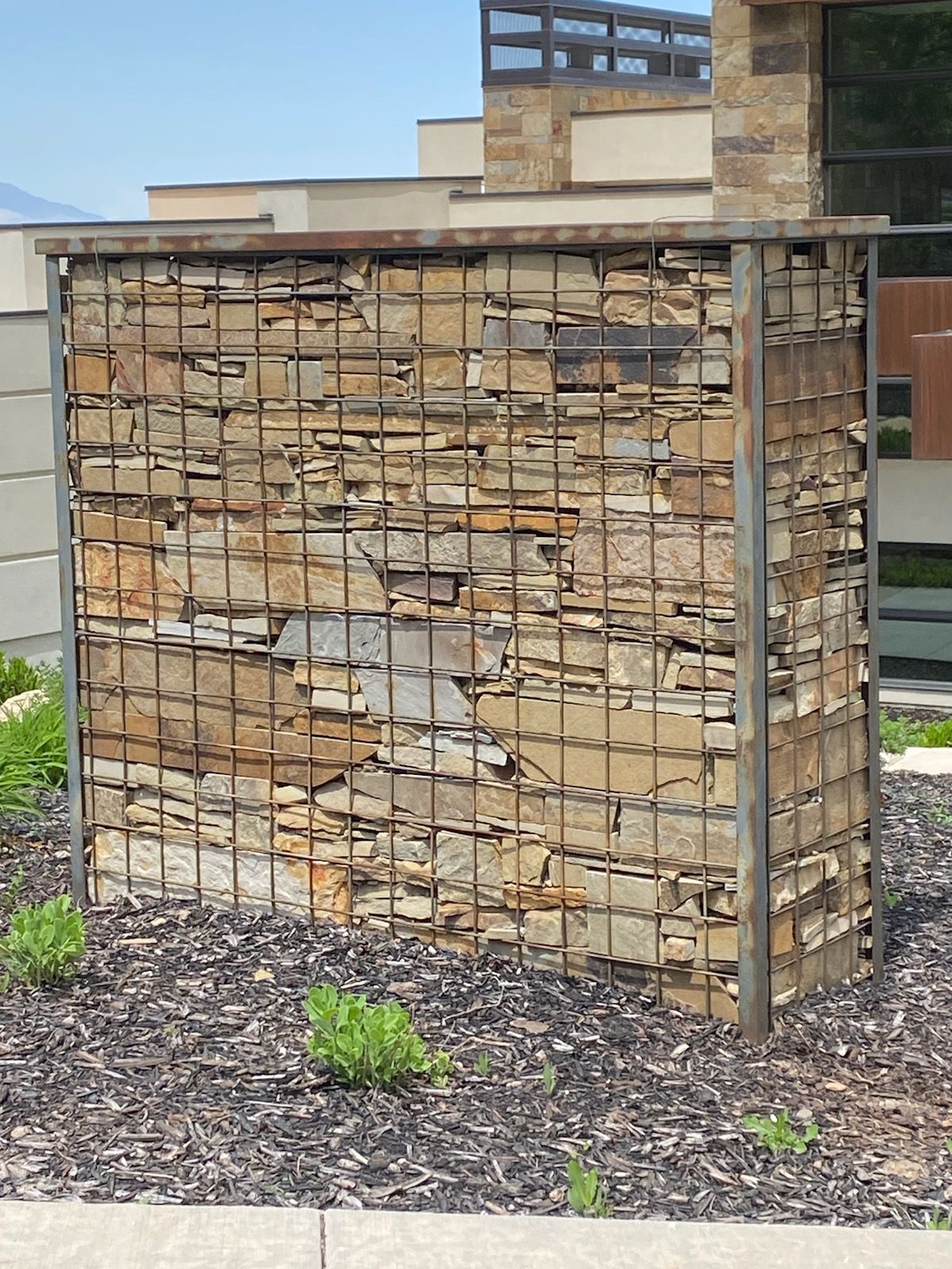 Gabion wall filled with brown, flat stones, framed by a metal grid, on a bed of dark mulch.