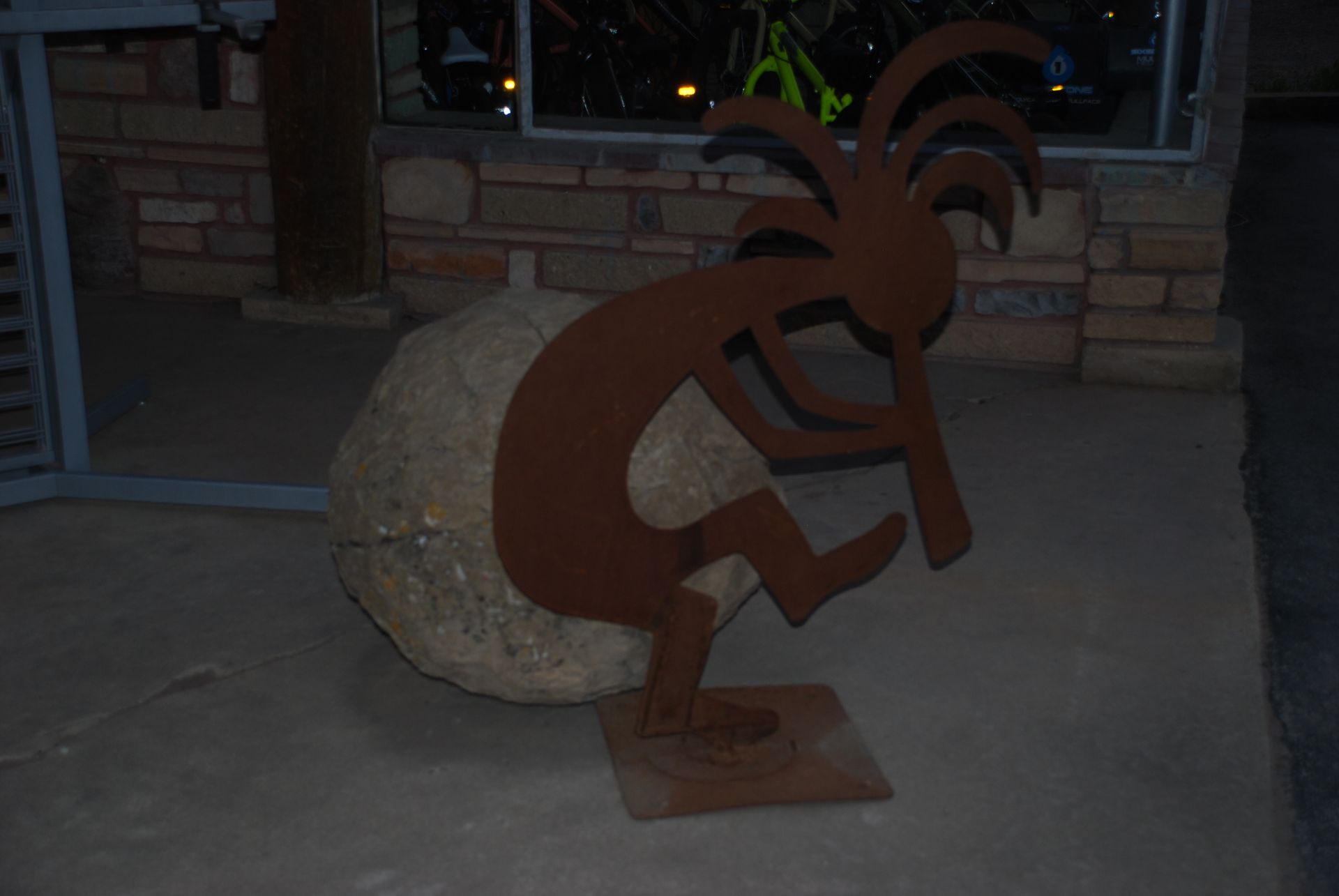 Metal Kokopelli sculpture leaning on a large rock on a concrete surface.