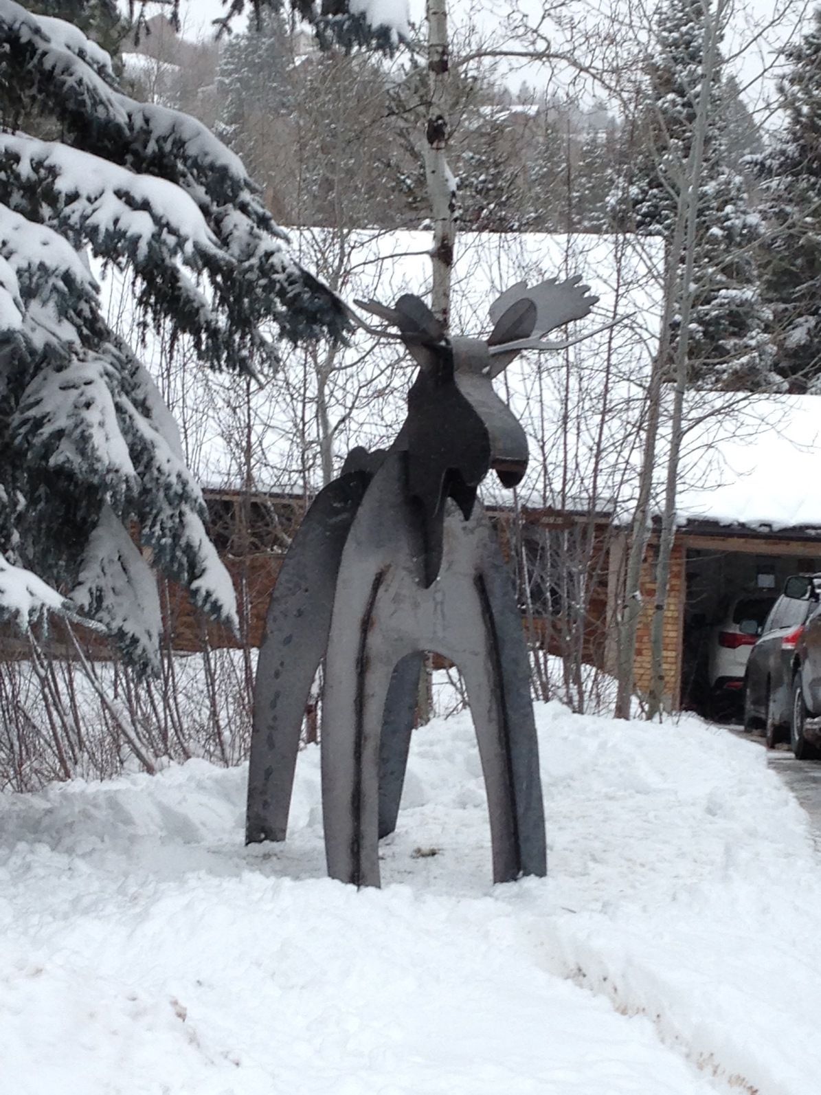 Metal moose sculpture in snow-covered yard with snowy trees and a building in the background.