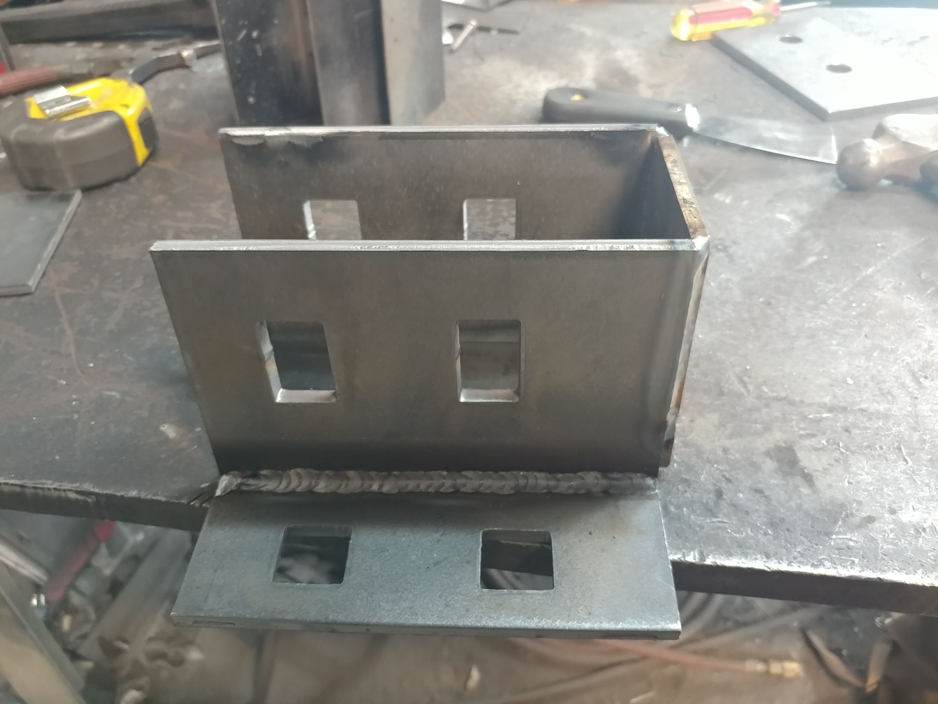Welded metal bracket with rectangular cutouts, sitting on a metal surface.