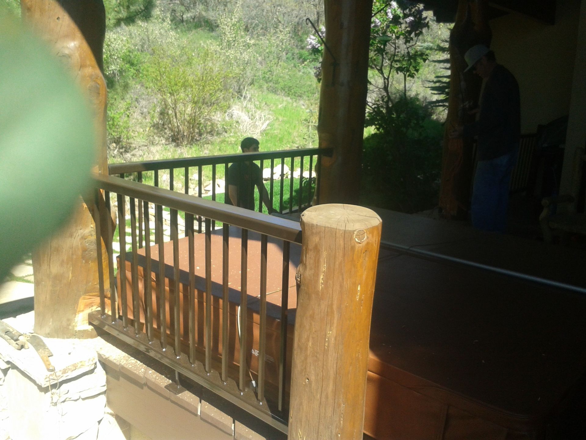 A porch with wooden railing and posts. Two people are visible: one outside, one inside, near the porch.