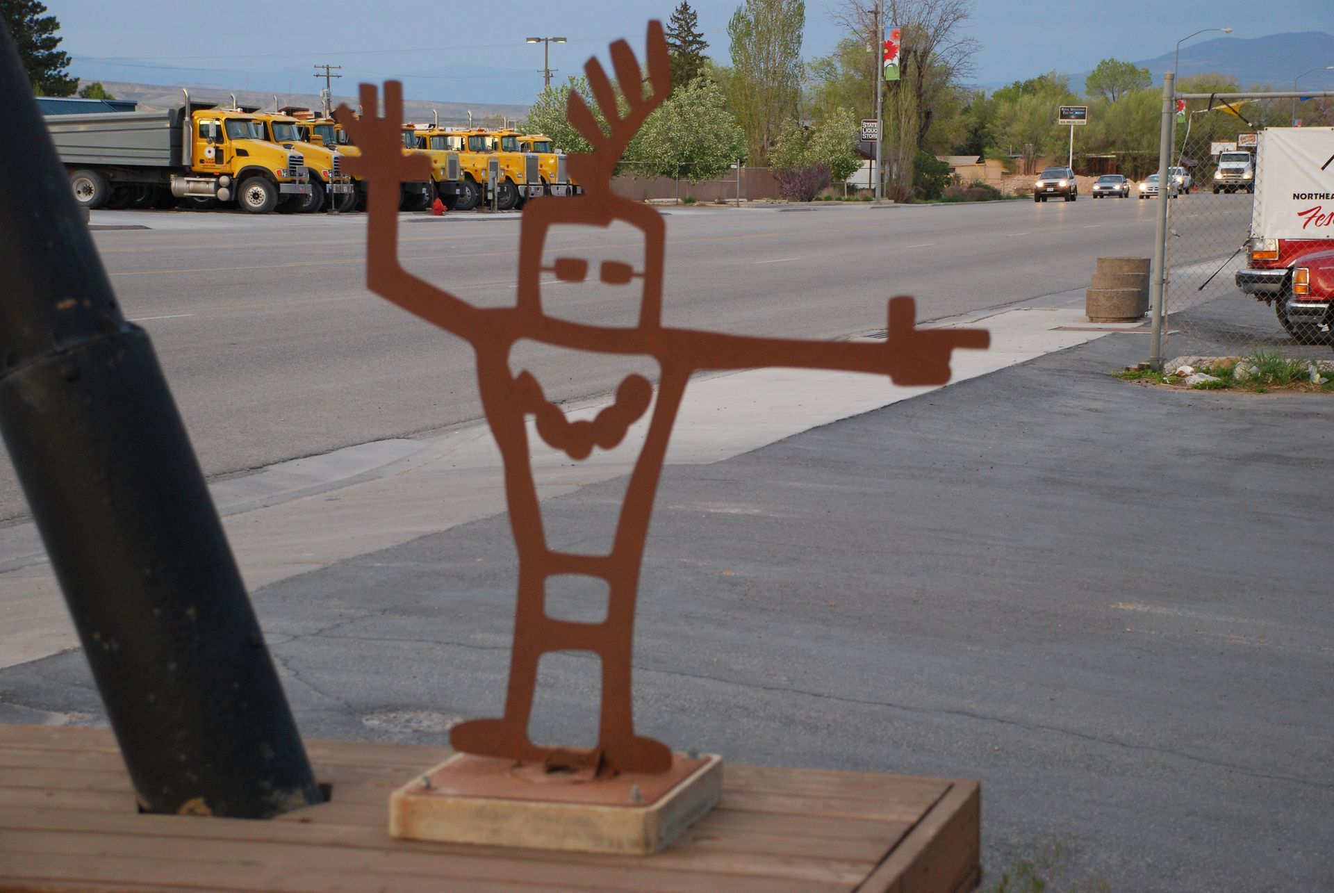 Rusty metal figure of a person with outstretched arms, standing outdoors; street and parked trucks in background.
