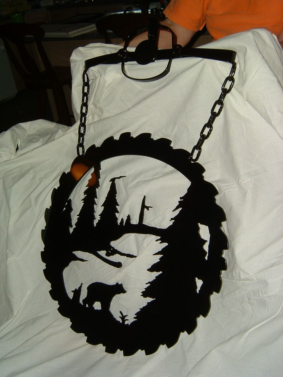 Black metal saw blade art with forest scene and bear, hanging by chains.