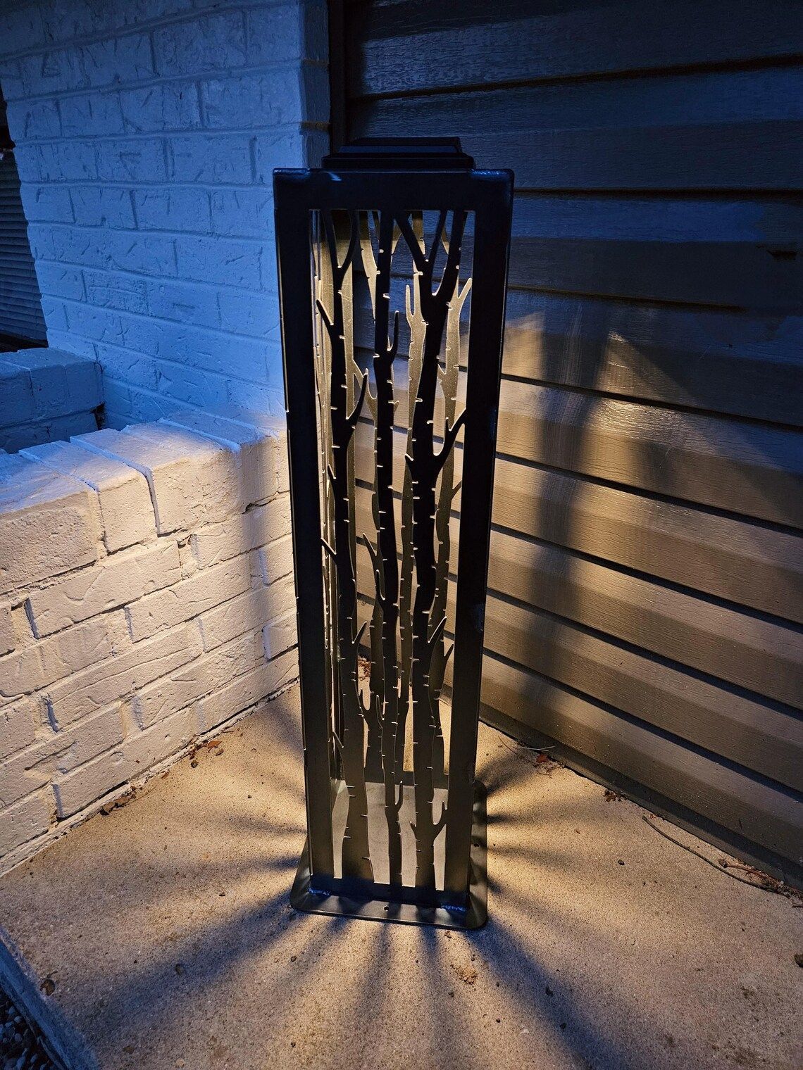 Black metal outdoor light with tree-branch design, casting light patterns on a concrete surface next to a brick wall.