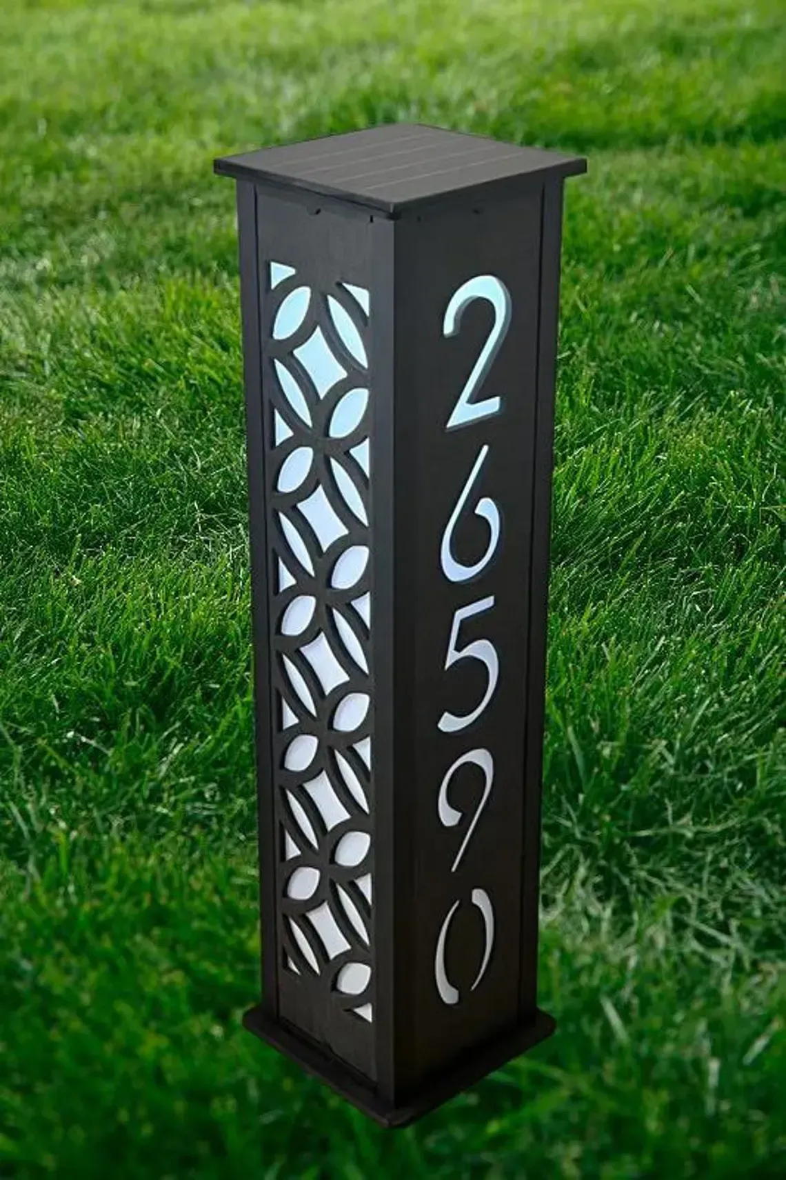 Dark brown illuminated address post with the numbers 26590 against a bright background, on green grass.