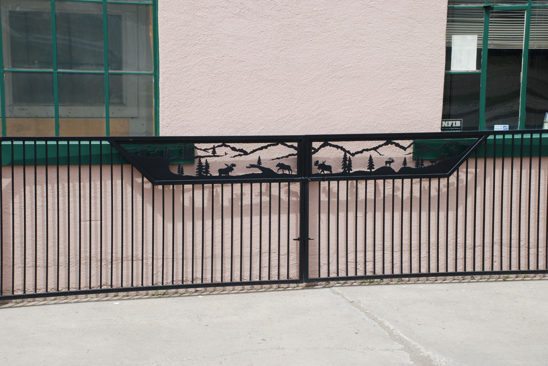 Black metal gate with a landscape design on top. Beige and green wall behind the gate.