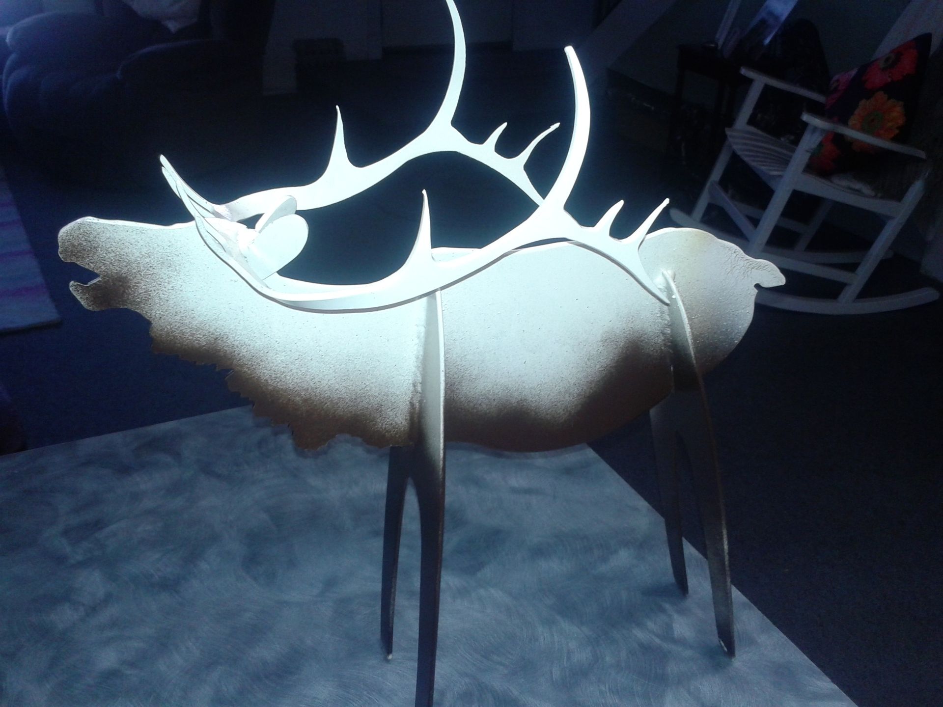 Wooden elk sculpture with large antlers and thin legs, standing on a textured surface.