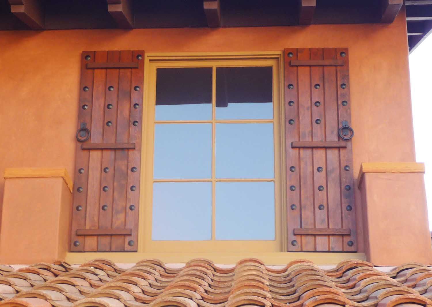 Window with wooden shutters on a terracotta-colored building, above a tiled roof.