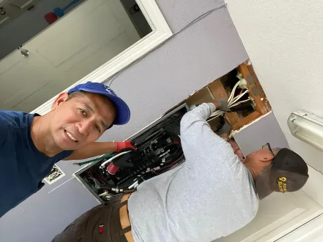 Two men working on electrical panel in a ceiling. One smiles at camera. Wires visible, room has a white door.