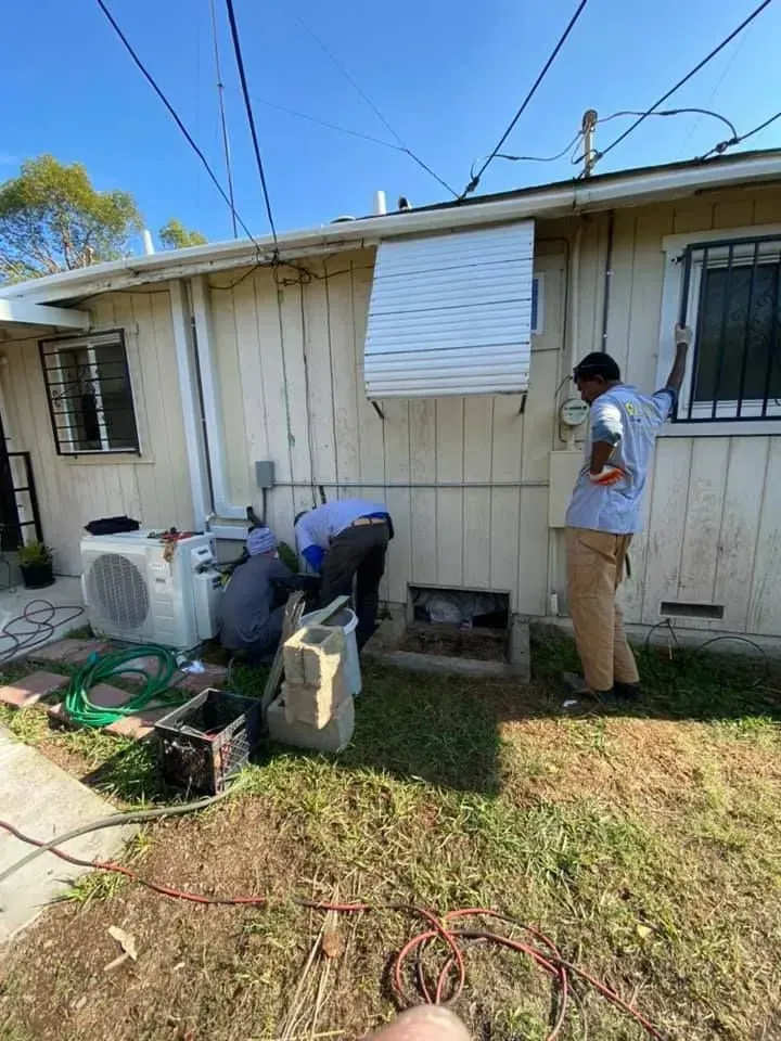 Three people working on the exterior of a building with electrical wiring visible, under a sunny sky.