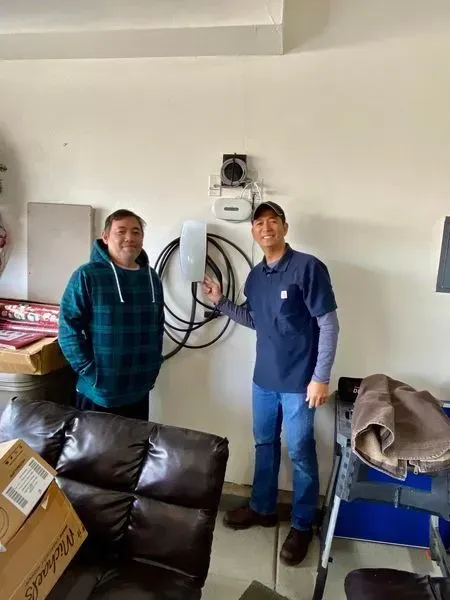 Two men in a garage beside an electric vehicle charger. One man points to the charger.