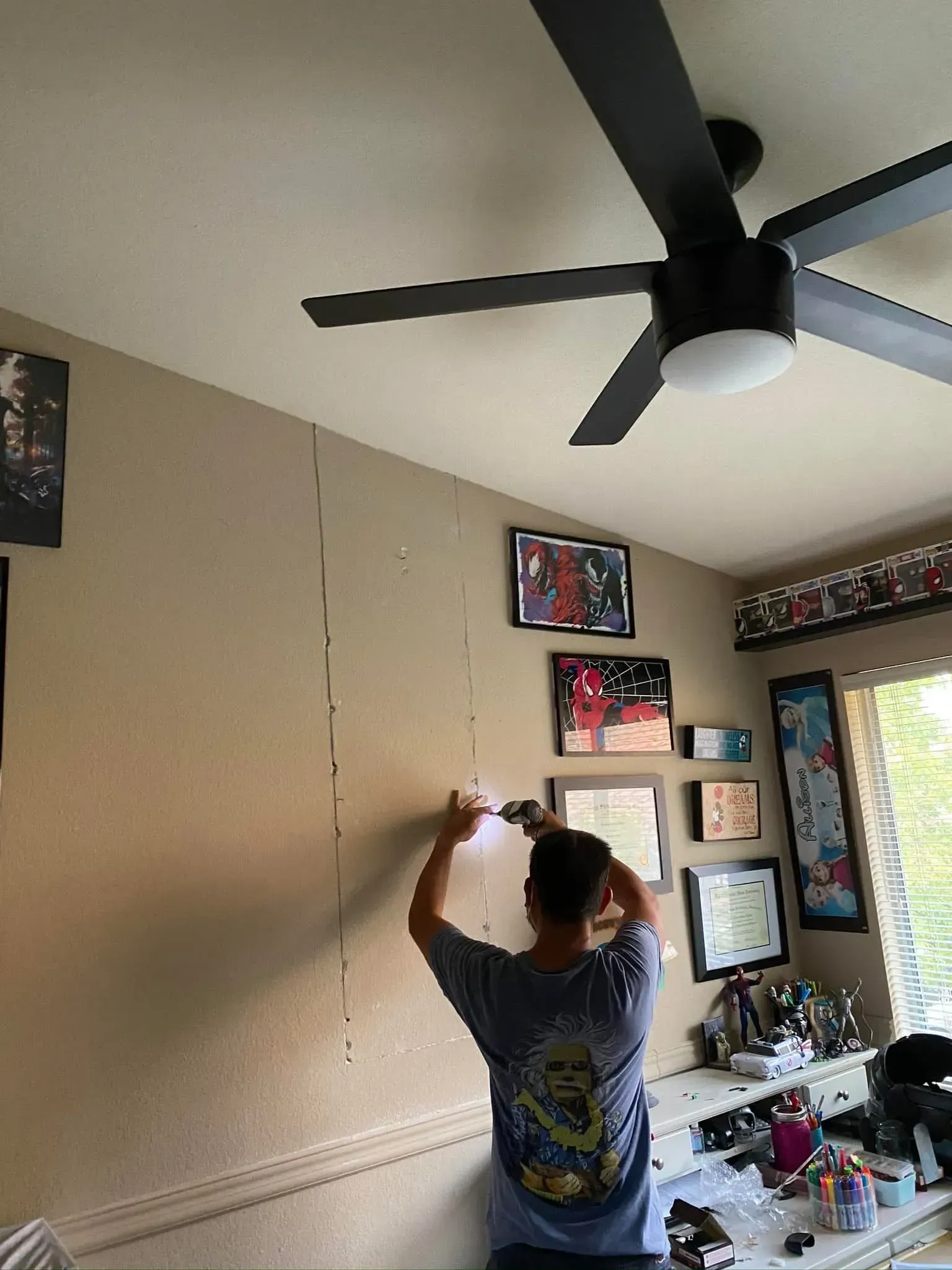 Person installing something on a tan wall with a power drill; ceiling fan and framed pictures nearby.