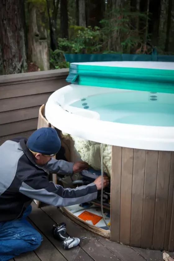 Person repairs hot tub on wooden deck, surrounded by trees.