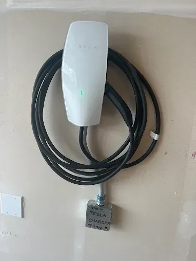White Tesla charger mounted on a beige wall, with a coiled black charging cable and a gray electrical box below.