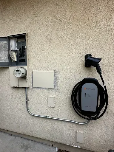 Electric vehicle charger mounted on stucco wall, with electrical panel and conduit visible.