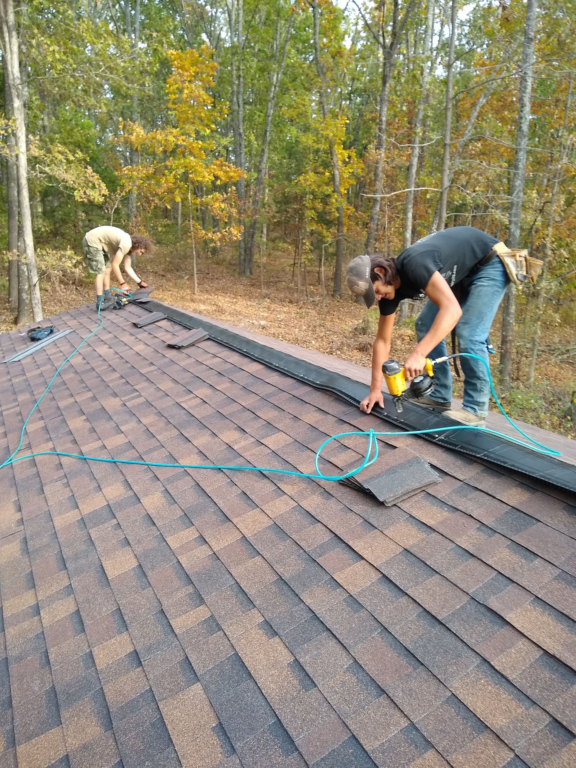 Two roofers working on a shingled roof in a wooded setting, one using a drill, the other laying shingles.