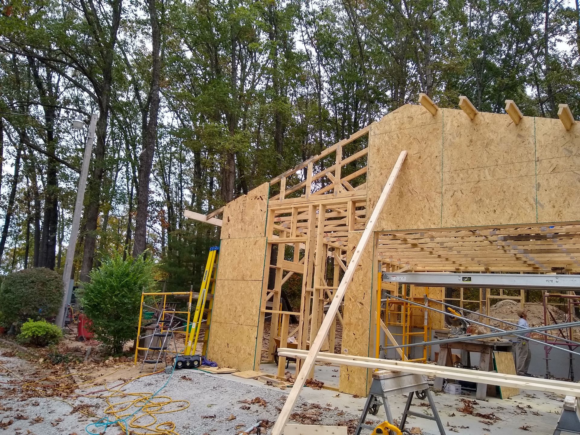 Construction of a wooden building with OSB siding, scaffolding, and a tall wooded background.