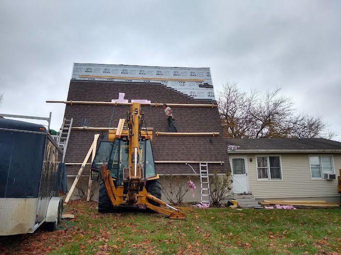 Construction workers on a roof installing shingles; a backhoe in front. Overcast day in a yard.