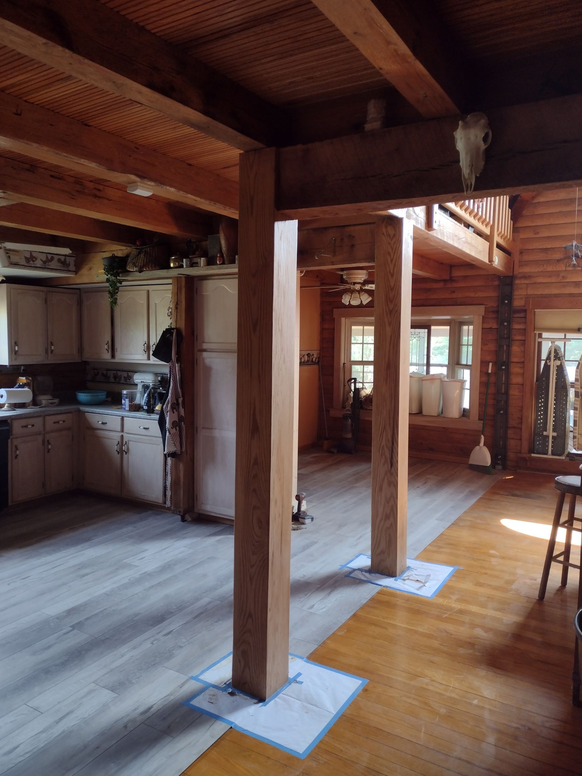 Two large wooden pillars in a rustic interior, supporting a second-story balcony. Kitchen is in the background.
