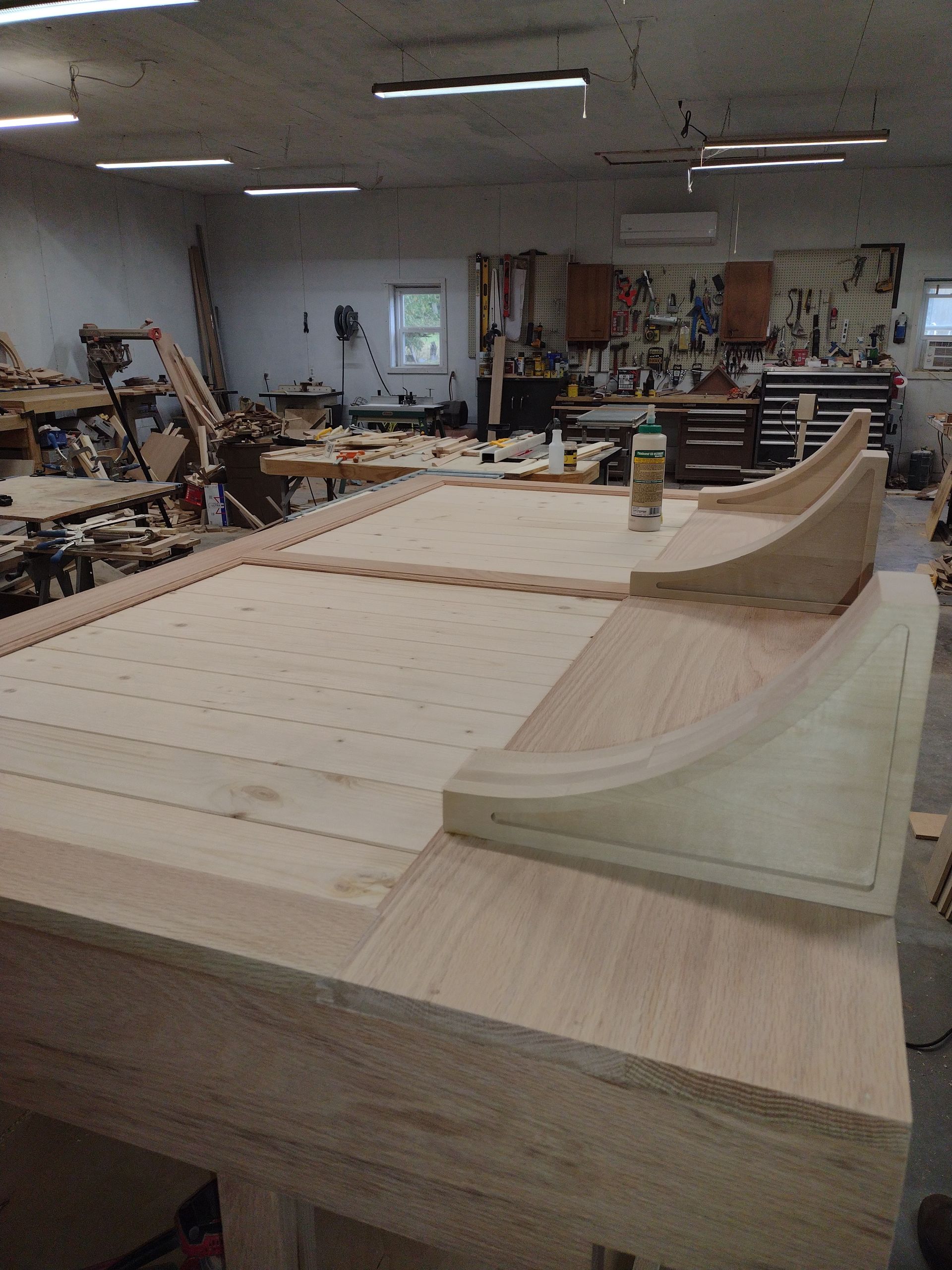 Wooden pool table under construction in a workshop. Light-colored wood, with tools and equipment visible in the background.