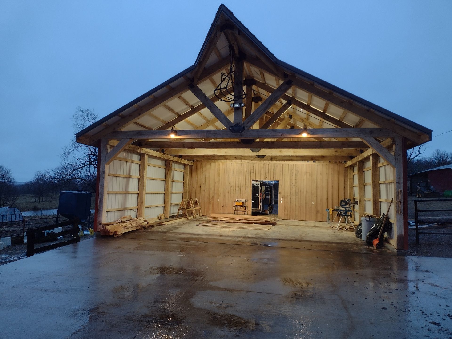 Open-air wooden barn with exposed beams, lit from within. Rain glistens on the ground.