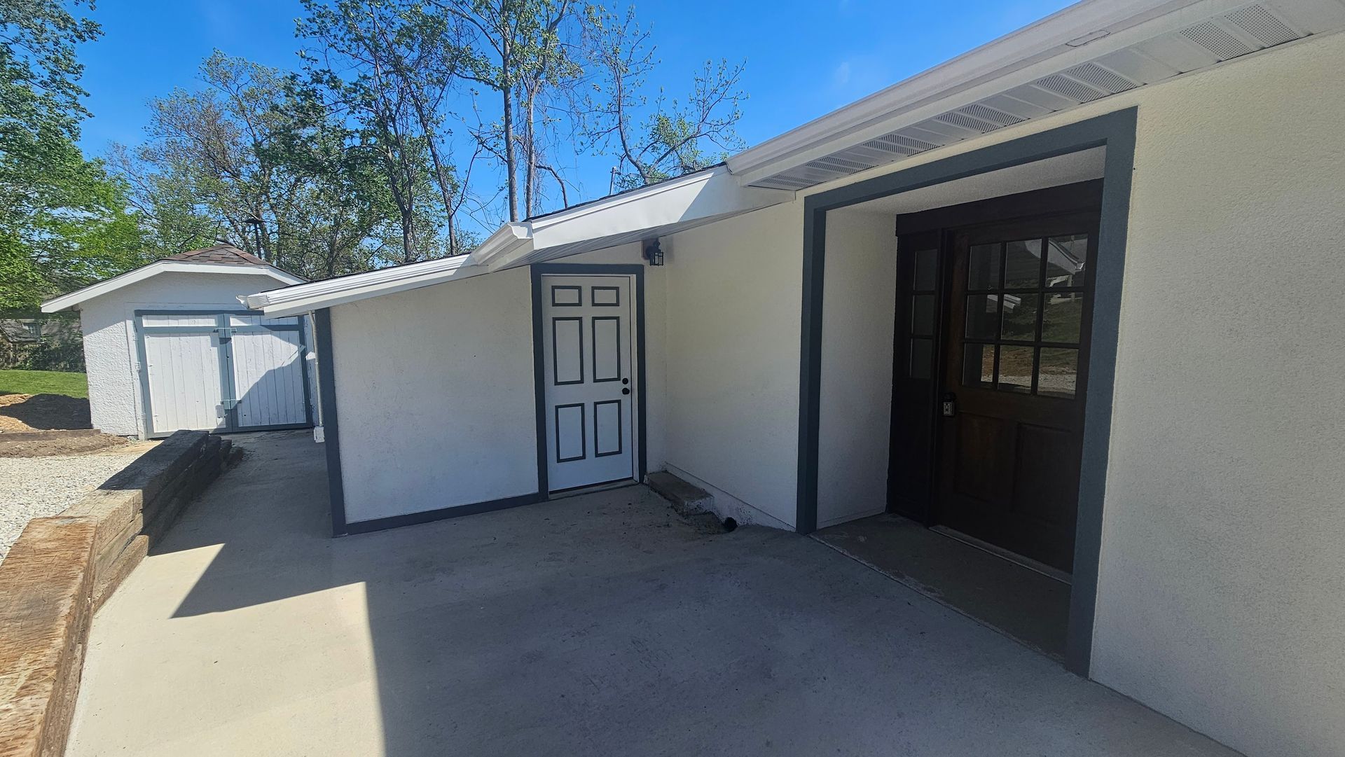 White building exterior with gray trim and a concrete patio; a shed is visible to the left.