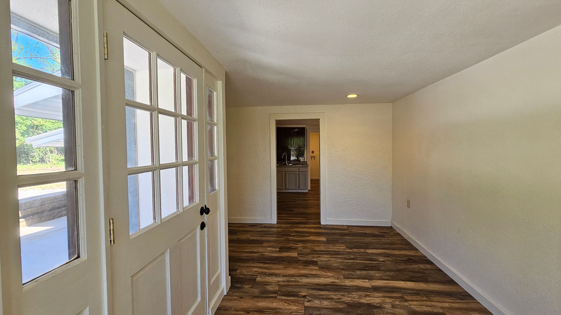 Entryway with open white French doors, leading to a porch. Brown floor and cream walls, leading to a kitchen.